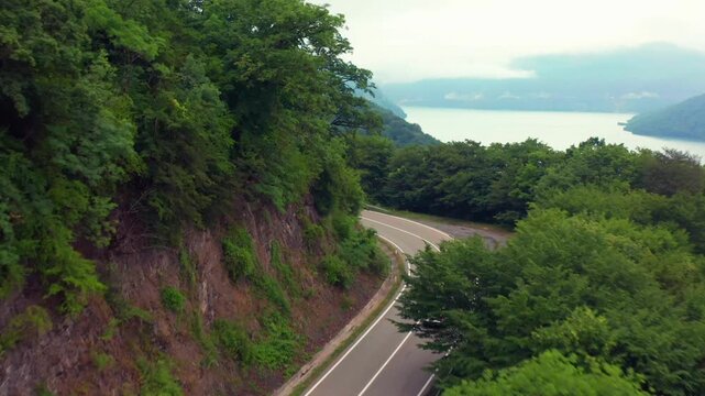 Aerial drone flies forward above a mountain road near Zhinvali Reservoir in Georgia, revealing blue water beyond the forested slope for scenic travel, tourism campaigns and regional transport media.