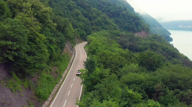 Aerial drone flies forward above a mountain highway near Zhinvali Reservoir in Georgia, following a car between forested slopes and open water for scenic driving, tourism access and travel media.