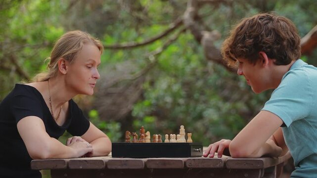 A mother and her teenage son play chess at a wooden picnic table outdoors in a park, concentrating on their strategy under green trees
