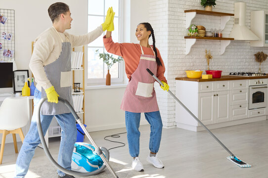 Couple cleaning home kitchen vacuuming mopping teamwork. Smiling man and woman high five while doing housework, using vacuum and mop on wooden floor. Teamwork and joyful home cleanliness.