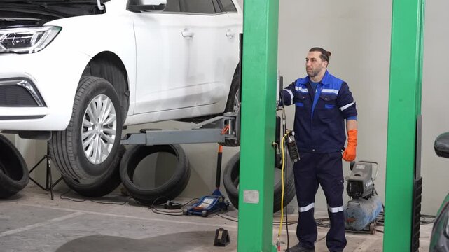 Auto repair mechanic in garage workshop performing vehicle maintenance on car lift, technician inspecting car on hydraulic hoist with tire tools spare wheels compressor, service repair scene