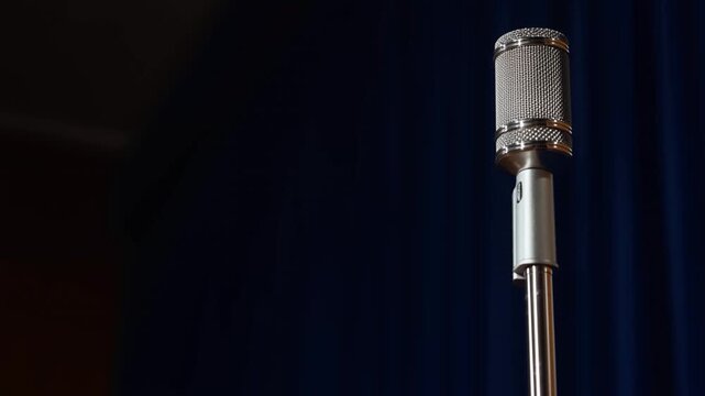 A steady, focused shot of a vintage silver microphone on a stand, set against a dark blue curtain backdrop in a professional studio or stage environment.