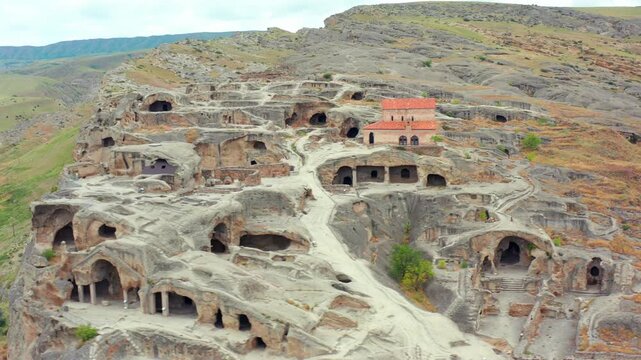 Drone circles beside Uplistsikhe cave complex in Georgia, framing rock-cut halls, tunnels and weathered terraces for heritage travel, ancient architecture research and tourism marketing campaigns.