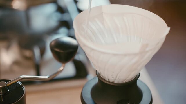 Pour over coffee brewing process, pouring hot water into ceramic dripper with paper filter, manual coffee grinder in background, specialty coffee preparation at home