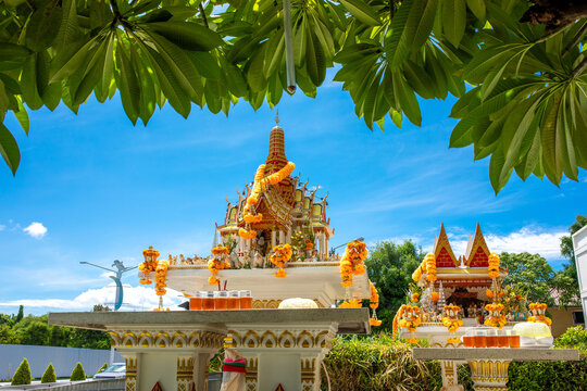 Traditional thai spirit house shrine decorated with flower garlands framed by green tropical tree leaves under blue sky