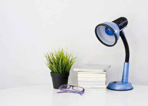 Minimalist workspace setup featuring desk lamp books glasses and small indoor plant on clean white surface