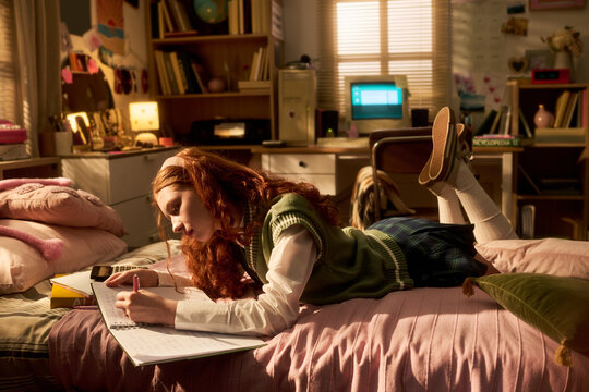 Teenage Caucasian girl studying on bed, writing in notebook in bedroom, preparing homework and exam revision. Education planning concept for school campaign, tutoring or teen learning content