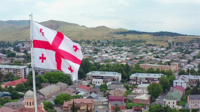 Georgian flag waves in aerial view over central Gori, historic roofs and surrounding hills, supporting civic identity, tourism advertising, regional culture and Georgian city branding campaigns.