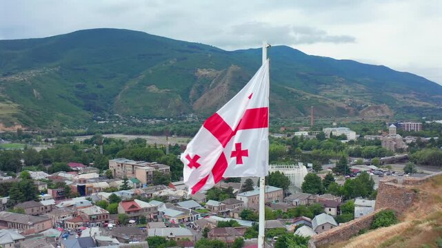 Georgian flag recedes in aerial view above Gori neighborhoods and green mountain slopes, supporting national branding, patriotic campaigns, travel promotion and regional identity media for Georgia.