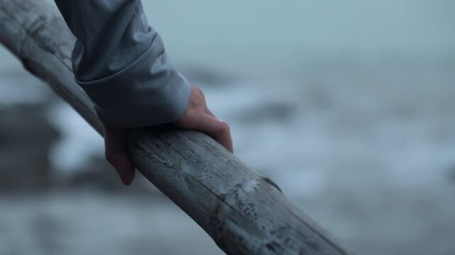 Hand grips rough old bamboo bridge pole by water's edge. Blurry waves at the background