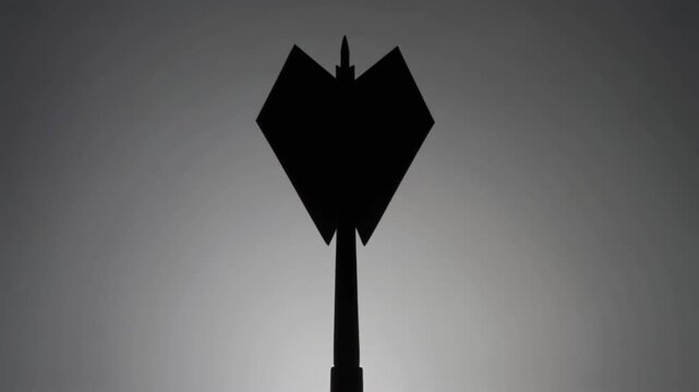 A dark silhouette of a dart-like object with stabilizing fins forming a heart shape, positioned vertically against a gradient gray background, symbolizing the contrast between love and conflict.