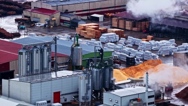 Aerial view of a sawmill facility with metal silos, stacks of timber, and industrial buildings in Latvia.