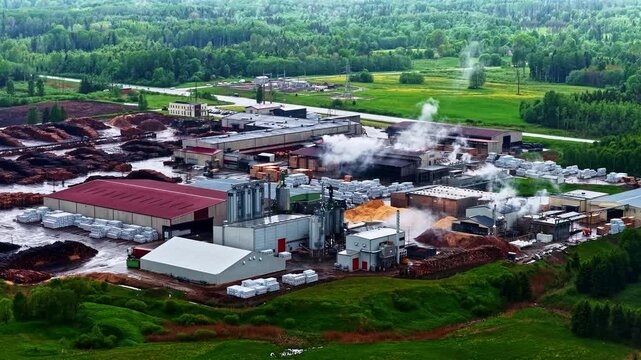 Aerial view of a wood processing plant with industrial buildings, steam rising from machinery, and large piles of timber surrounded by lush green forests in Latvia.
