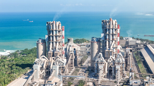 Aerial view of a large industrial cement factory complex with tall silos and processing towers situated along a lush green coastline overlooking the blue sea in Indonesia.