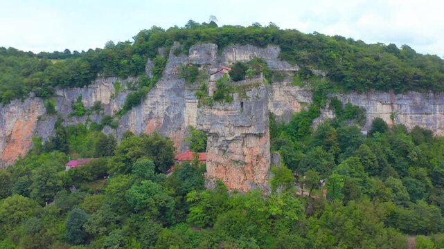Aerial drone sweeps sideways around Katskhi Pillar in Georgia, revealing the monolith beneath forested cliffs for pilgrimage tourism, religious heritage, conservation and regional travel media.