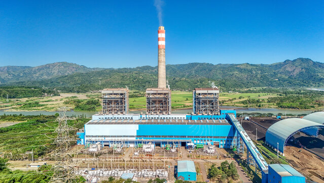 Aerial view of a large power plant with a tall chimney emitting smoke, blue industrial buildings, and surrounding green hills in Indonesia.