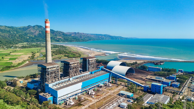 Aerial view of a large power plant with a tall chimney and industrial facilities situated along the coastline near green hills and the blue ocean in Indonesia.