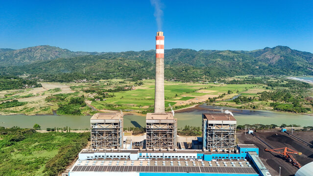 Aerial view of a power plant with a tall chimney and industrial buildings situated near a river and lush green mountains in Indonesia.