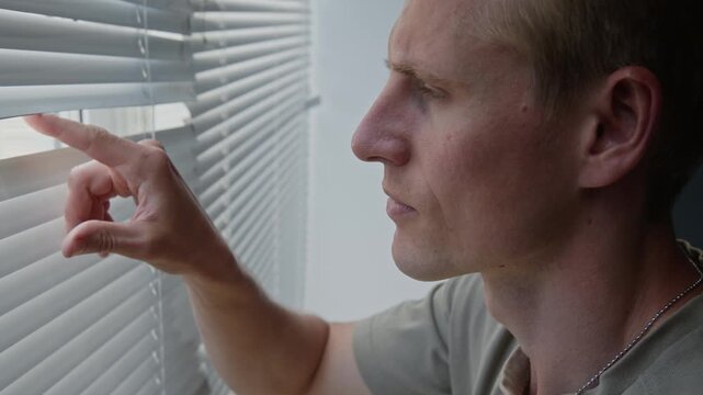 Caucasian young adult man wearing dog tags looking through window blinds with serious expression. Footage for mental health awareness campaigns, PTSD support, or security services