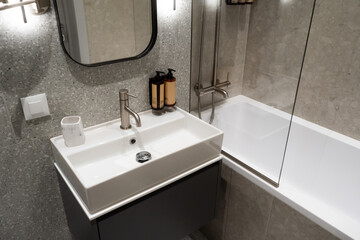Fototapeta premium Contemporary bathroom featuring a stylish vanity sink, sleek faucet, and white bathtub, surrounded by gray terrazzo and large format tiles, reflecting modern design and comfort