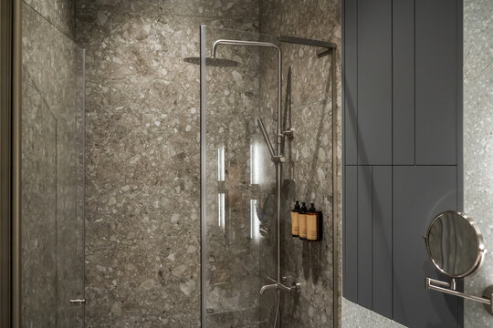 Contemporary bathroom interior featuring a walk in shower with terrazzo tiled walls, a glass enclosure, rain shower head, and dark grey minimalist storage solutions