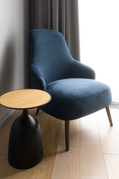 Blue velvet armchair standing next to a modern side table with a wooden top and black base, furnishing a contemporary room interior with a wood floor and curtains