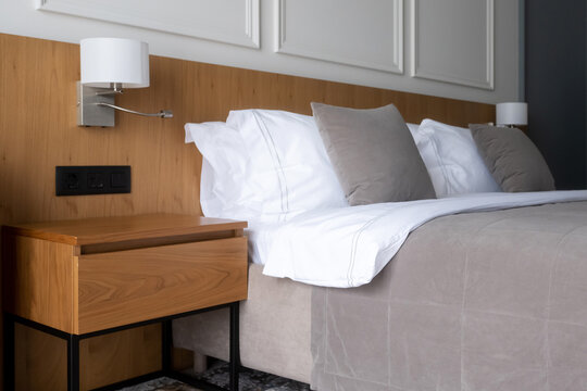 Modern hotel room showcasing clean white and neutral toned bedding on a bed with a wooden headboard, complemented by a contemporary wooden bedside table with a sleek lamp and power outlets