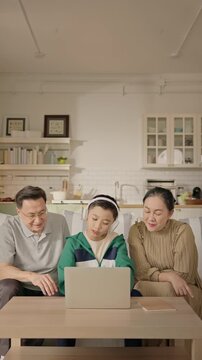 Asian boy sits on sofa with his grandparents, patiently showing them how to use a laptop in cozy home, intergenerational learning, digital literacy for seniors, family connection across generations.