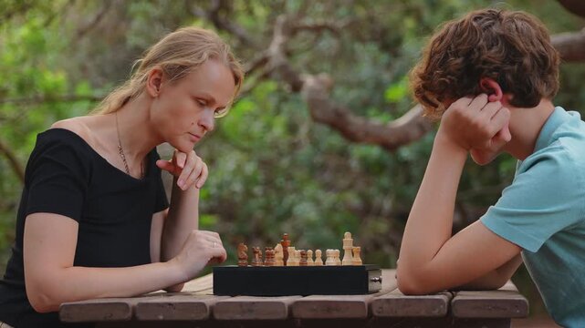 A mother and her fifteen year old teenage son concentrate, as they play a game of chess on a wooden picnic table outdoors in a green park