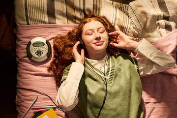 Teenager girl lying on bed, listening through headphones beside portable CD player, relaxing during study break and expressing youth leisure. Top view, ideal for teen lifestyle campaigns © pressmaster