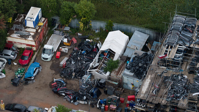 Dagenham, United Kingdom - 10 July 2026: Aerial view of a car salvage yard filled with piles of automotive parts, scrap metal, and stacked bumpers on scaffolding, surrounded by industrial fencing.