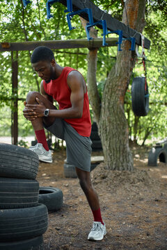 Vertical shot of young Black man stretching knee on tire during outdoor obstacle workout, preparing for fitness training and endurance challenge. Ideal for sports wellness campaigns