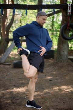Vertical shot of young adult Caucasian man stretching quadriceps during outdoor obstacle workout, preparing body for fitness training and injury prevention. Useful for sports wellness marketing
