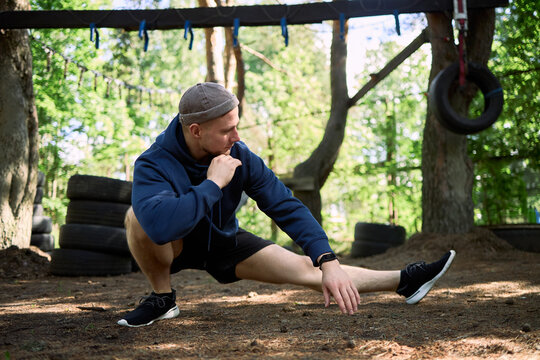 Young adult Caucasian man stretching legs during outdoor obstacle course workout, preparing for fitness training and mobility practice. Useful for sports wellness marketing and gym promotion