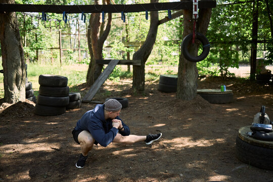 Wide shot of young adult man stretching legs during outdoor obstacle course workout, preparing for fitness training and endurance challenge. Useful for sports marketing and wellness campaigns