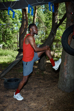 Vertical shot of young Black man stretching leg against tree while warming up at outdoor obstacle course, preparing for fitness training. For sports wellness, endurance, active lifestyle marketing