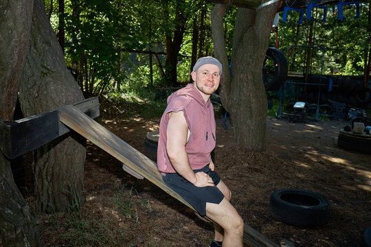 Portrait of young adult Caucasian man sitting on outdoor obstacle course equipment, smiling at camera in fitness park. Suitable for workout promotion, wellness branding, medium shot