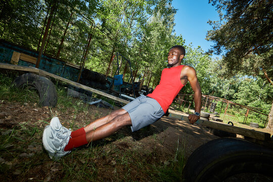 Young Black man exercising on outdoor obstacle course, performing tricep dips on wooden beam in forest setting, showing fitness training and endurance. Useful for sports marketing