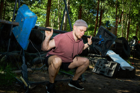 Caucasian young adult man squatting with barbell in outdoor gym, training lower body strength and endurance. Fitness motivation concept for workout programs, sports coaching, healthy lifestyle