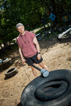 Vertical shot of young adult Caucasian man training outdoors, striking tractor tire with sledgehammer during functional workout. Fitness motivation concept for gym promotion or sports ads