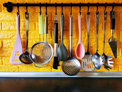 Bright kitchen utensils hanging on a railing against a yellow brick wall&mdash;a spatula, a slotted spoon, tweezers, a ladle, a whisk, a sieve, and a spoon. Silicone and metal utensils