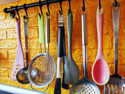 Bright kitchen utensils hanging on a railing against a yellow brick wall&mdash;a spatula, a slotted spoon, tweezers, a ladle, a whisk, a sieve, and a spoon. Silicone and metal utensils