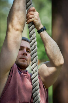 Vertical shot of Caucasian young adult man climbing rope during outdoor obstacle training, showing strength and endurance. Ideal for fitness marketing, boot camp promotion, wellness content