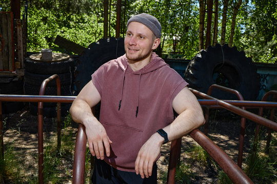 Medium shot of young adult Caucasian man resting between outdoor workout bars, smiling away from camera during fitness training. For gym promotion, wellness branding, active lifestyle marketing
