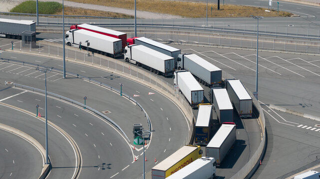 Calais, France - 09 July 2026: Aerial view of a long queue of semi-trucks waiting to enter the Port of Calais ferry terminal, with white trailers lined up along the curved road.
