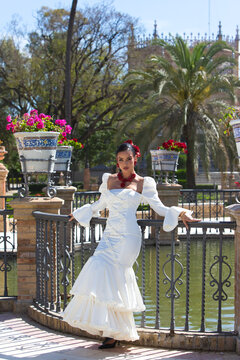 A beautiful woman dancing flamenco in a park in Seville, Spain. She is wearing a traditional white dress with ruffles and has red flowers in her hair. Flamenco, a UNESCO Intangible Cultural Heritage.