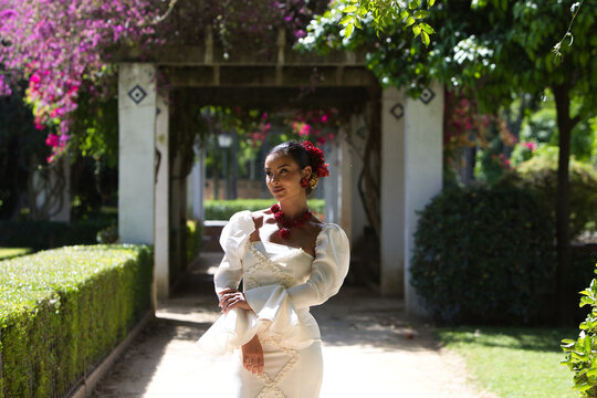 A beautiful woman dancing flamenco in a park in Seville, Spain. She is wearing a traditional white dress with ruffles and has red flowers in her hair. Flamenco, a UNESCO Intangible Cultural Heritage.
