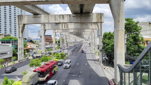 Elevated street view of a major multi-lane road in Bangkok, Thailand, framed by large concrete pillars of the skytrain infrastructure with cars and a red bus driving by.