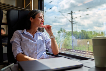 Woman passenger in a train looking out of the window, seeing countryside during her commute, reflecting and thinking while enjoying the journey with a laptop and coffee © DusanJelicic