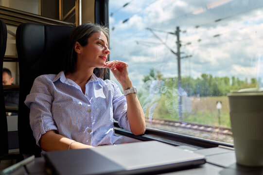 Woman passenger in a train looking out of the window, seeing countryside during her commute, reflecting and thinking while enjoying the journey with a laptop and coffee